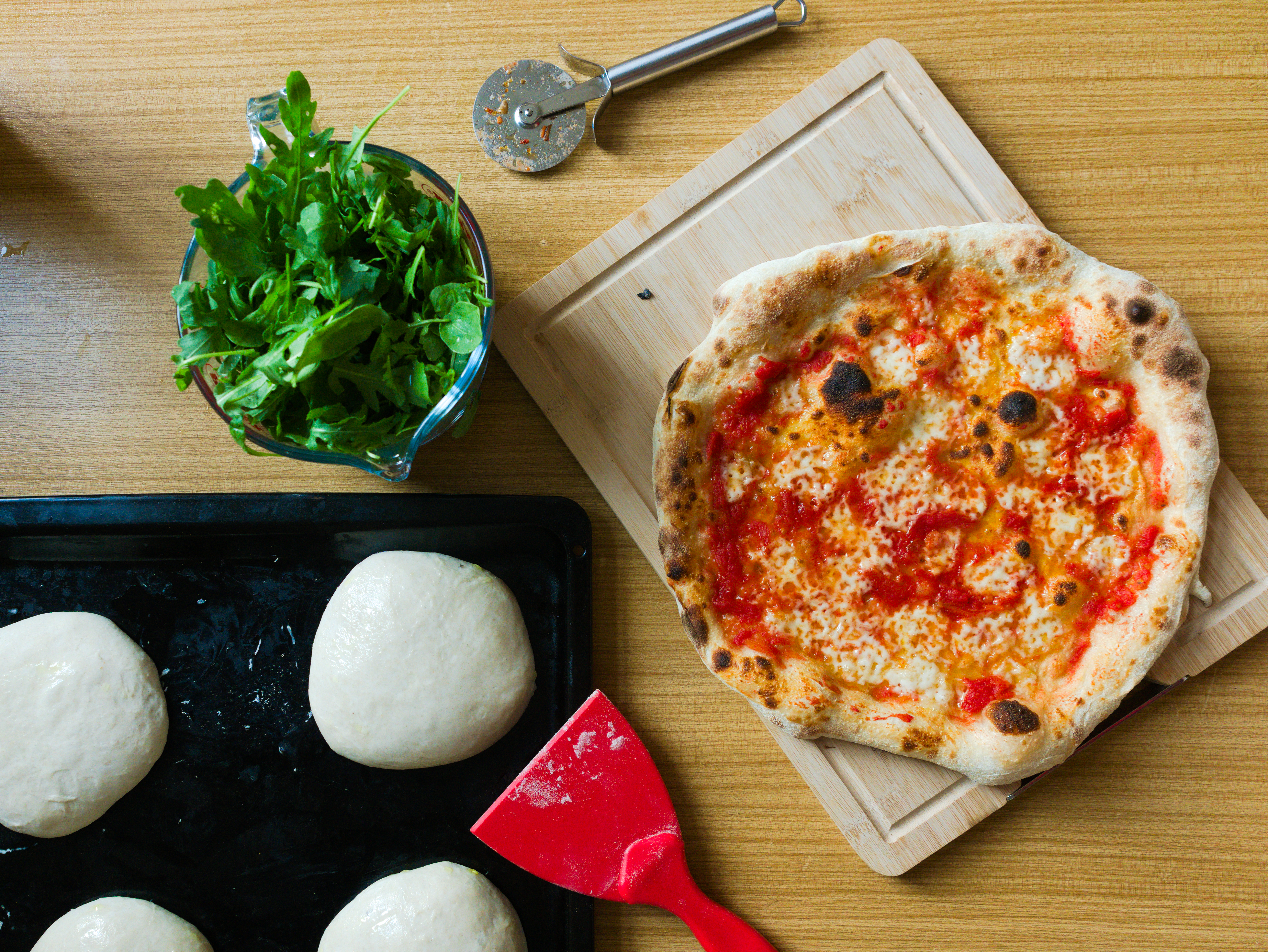 Eine rote Pizzakarte, ein Pizza-Teigling, grüner Rucola und eine fertig gebackenen neapolitanische Pizza liegen auf einem Tisch.